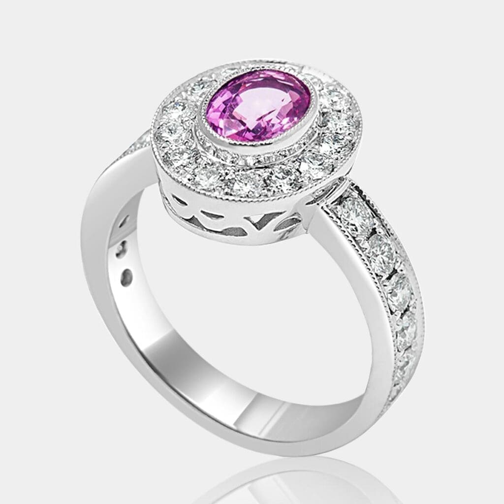 Handmade, designer style engagement ring featuring oval cut pink sapphire and halo of round diamonds, with diamonds in the band and milgrain detail.